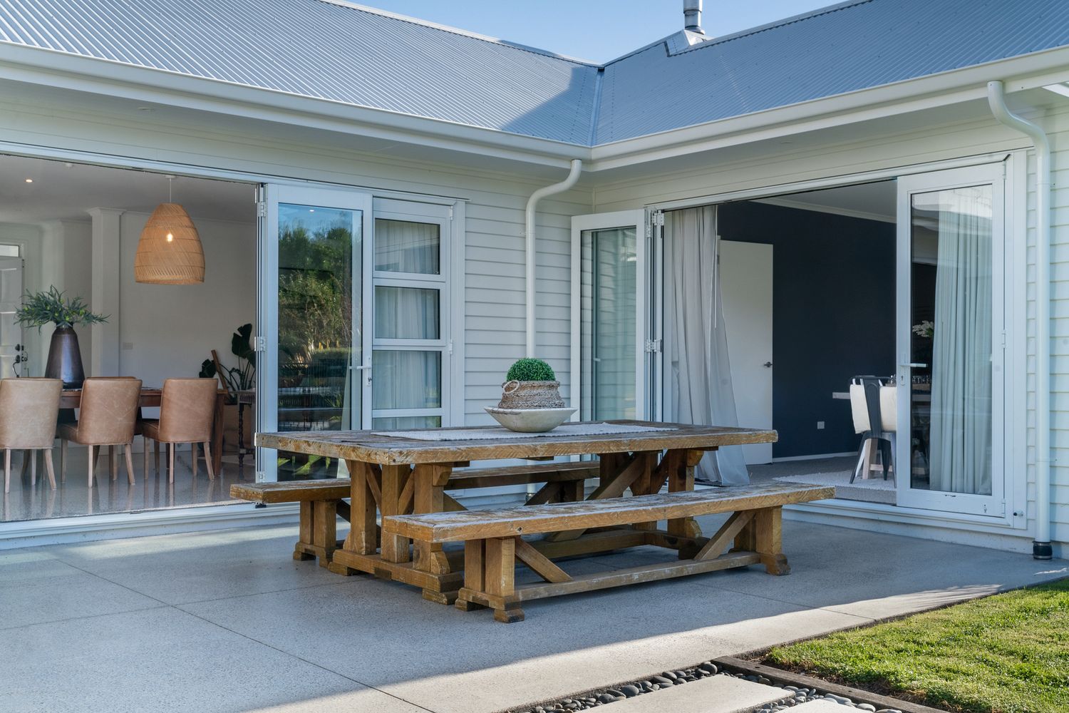 Modern outdoor patio area with wooden picnic table and dining area connected to white weatherboard house.