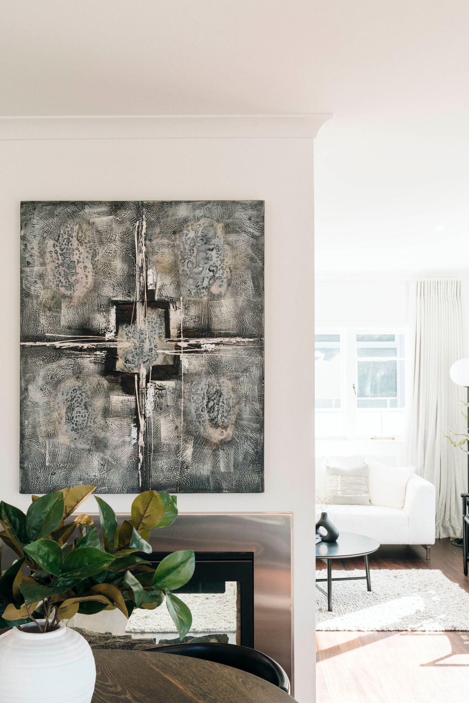 Large abstract grey cross painting above green plants in a modern white living room.
