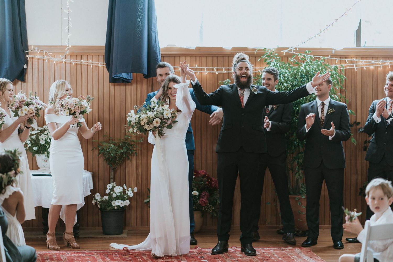 Wedding guests dance and celebrate together at reception in wood-paneled venue with festive party atmosphere.