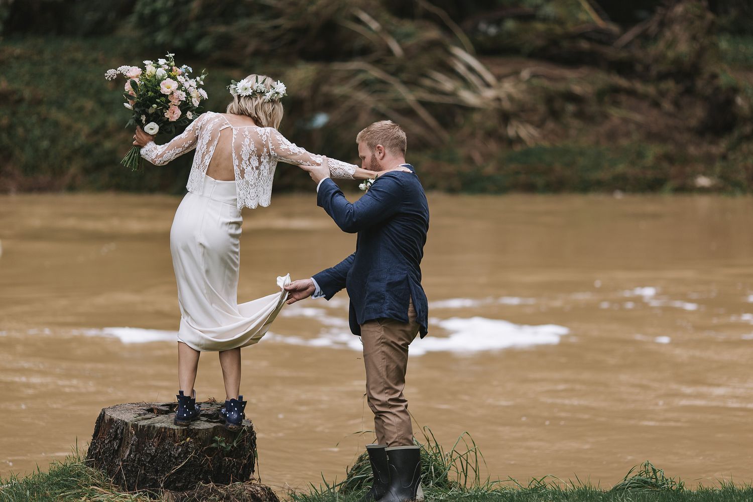 A couple in wedding attire stands on mossy rocks beside a muddy river during their outdoor celebration.