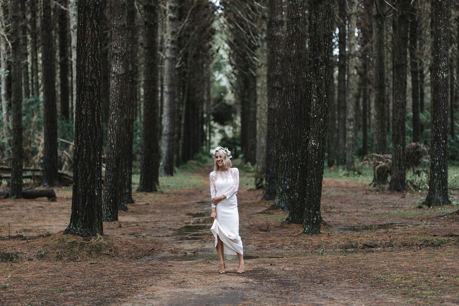 A graceful figure in flowing white dress stands contemplatively among tall evergreen trees in a serene forest setting.