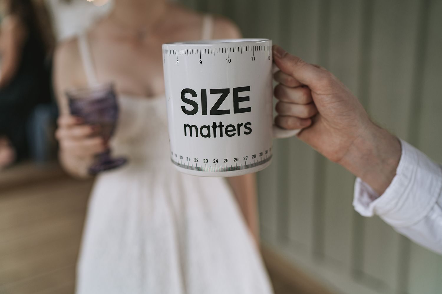 A hand holds a white coffee mug with measuring tape design and text reading 'SIZE matters' against a blurred background.