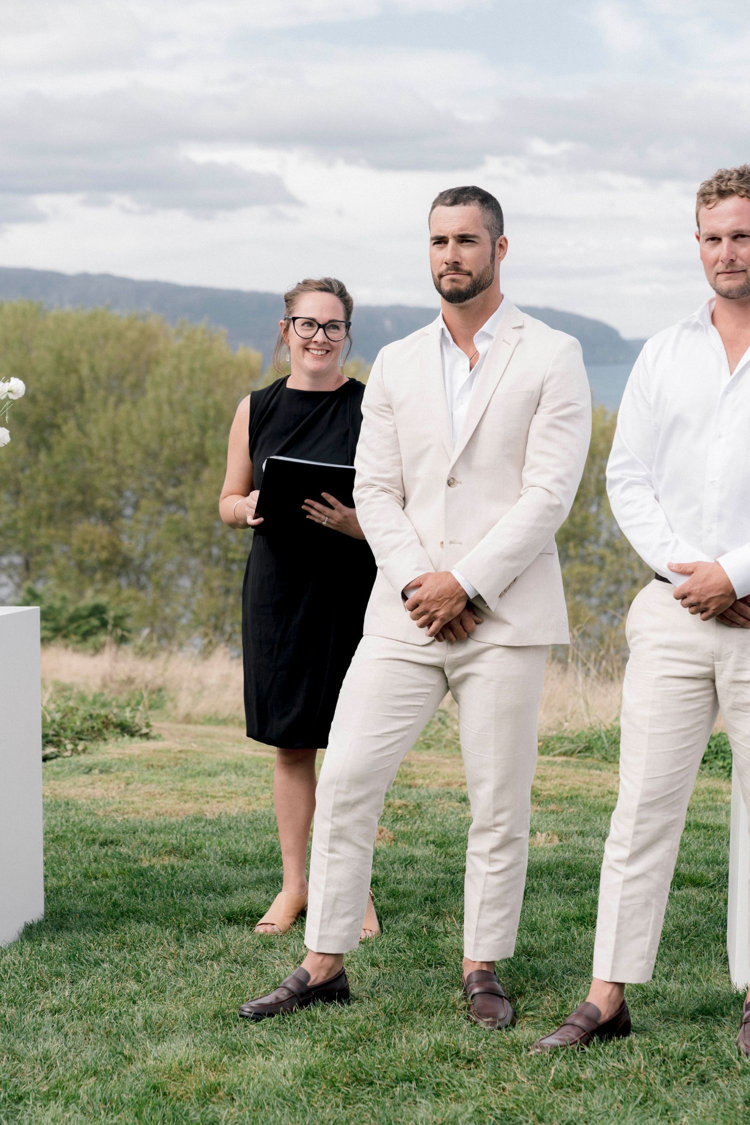 Wedding ceremony setup outdoors with officiant and groomsmen against mountain backdrop.