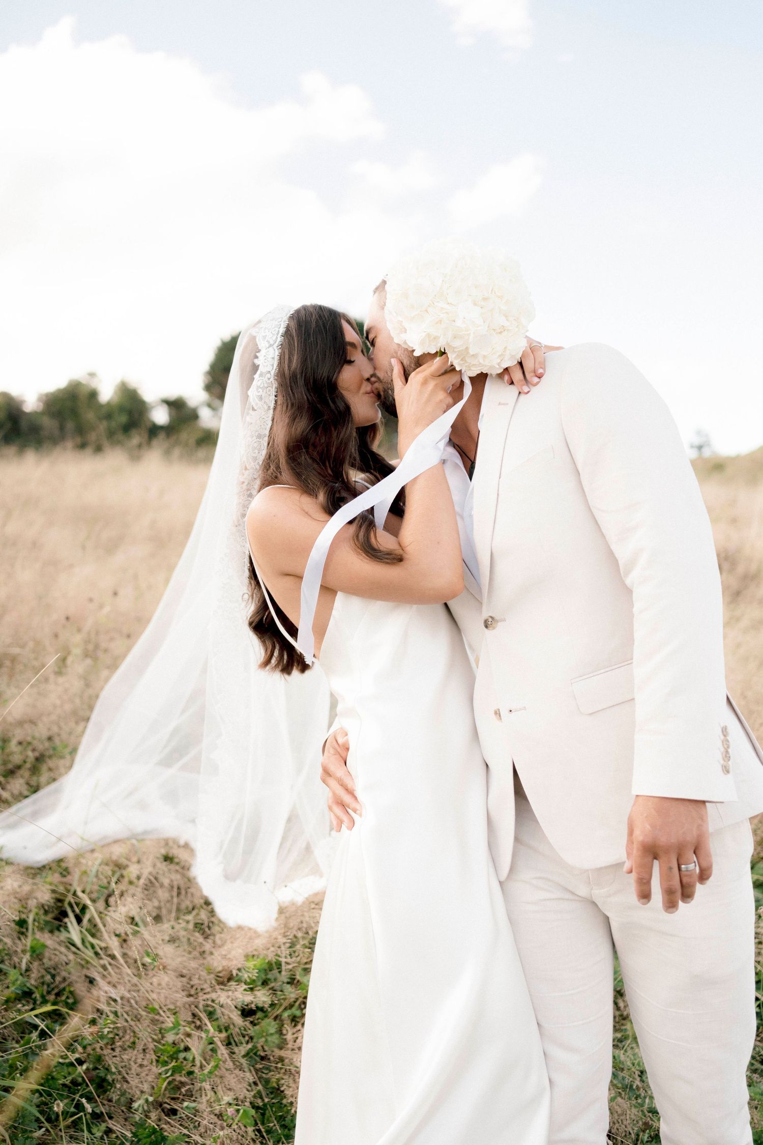 A couple in wedding attire share a romantic moment in a sunlit field with flowing white dress and veil.