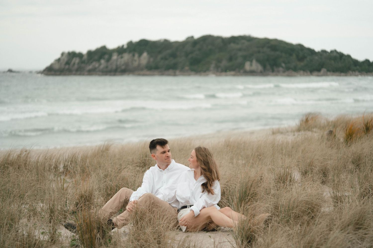 A couple embraces and relaxes in tall beach grass beside the ocean on a cloudy day.