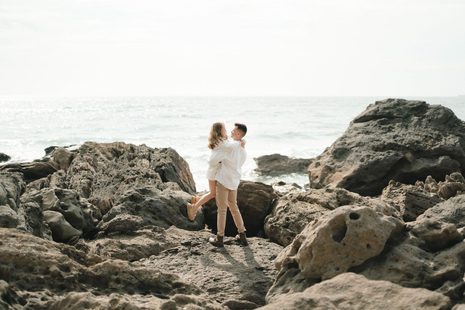 Couple exploring scenic rocky coastline with waves crashing against volcanic rocks at sunset.