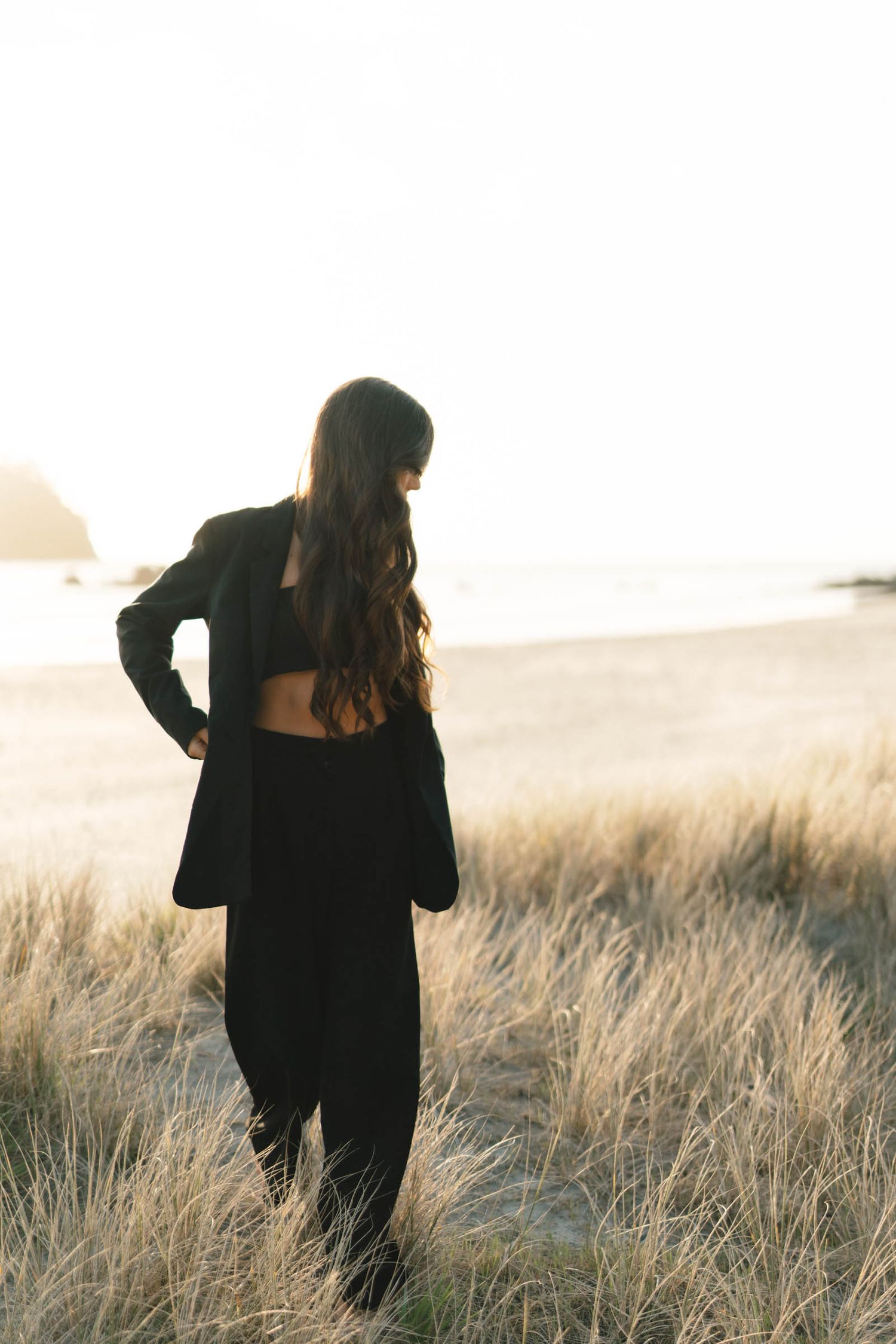 Person wearing black clothing stands among tall grass on coastal dunes at sunset.