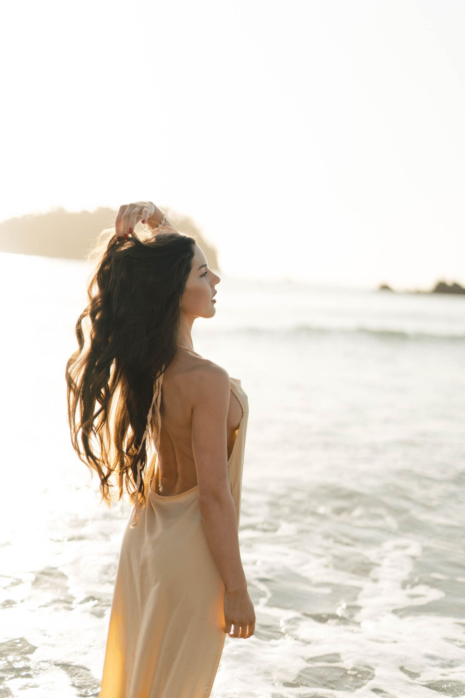 Sunlit beach portrait of a person in a light dress with windswept hair against sparkling ocean waves.
