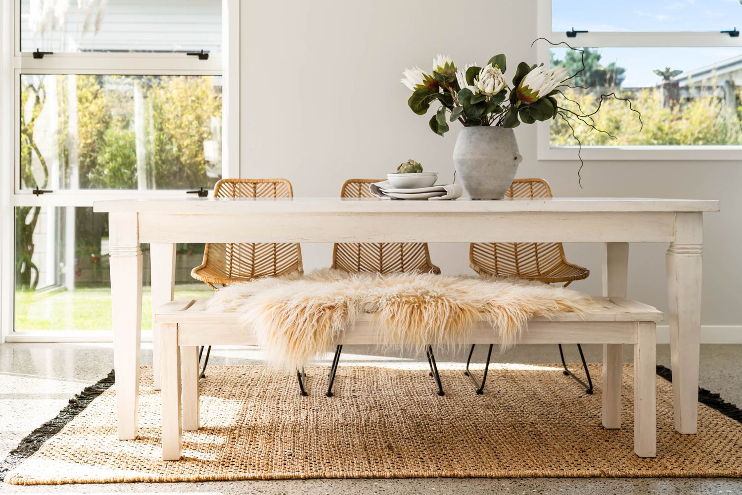 Bright coastal-style dining area with jute rug, white furniture and large windows overlooking green outdoor space.