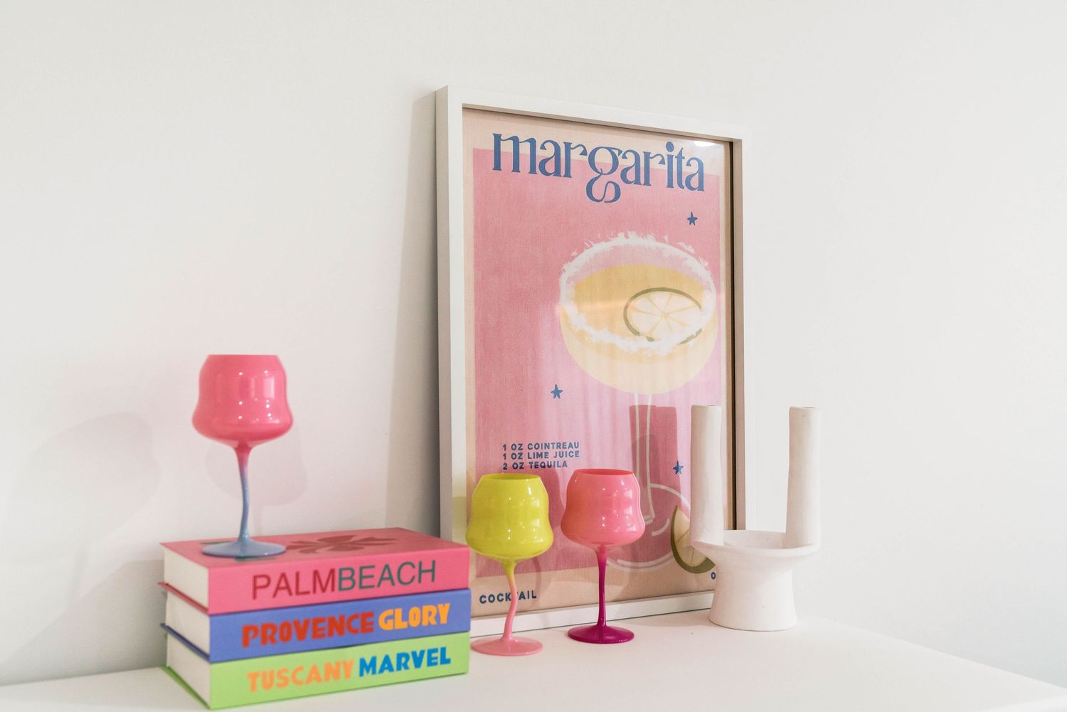 Pink wine glasses and cocktail books arranged on white shelf with soft pastel decor accents create a stylish bar display.