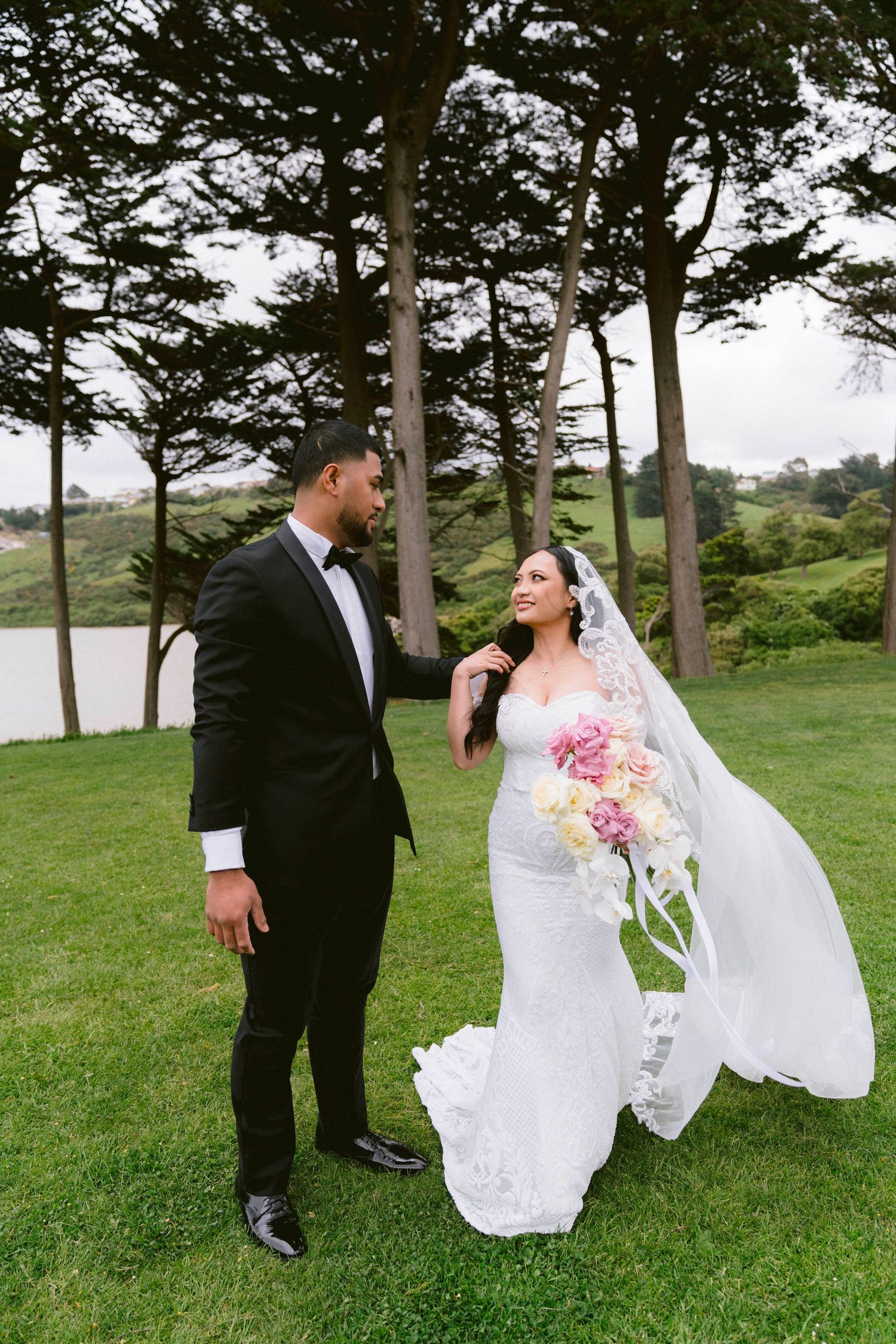 Newlywed couple shares a romantic moment outdoors near a lake surrounded by tall trees.