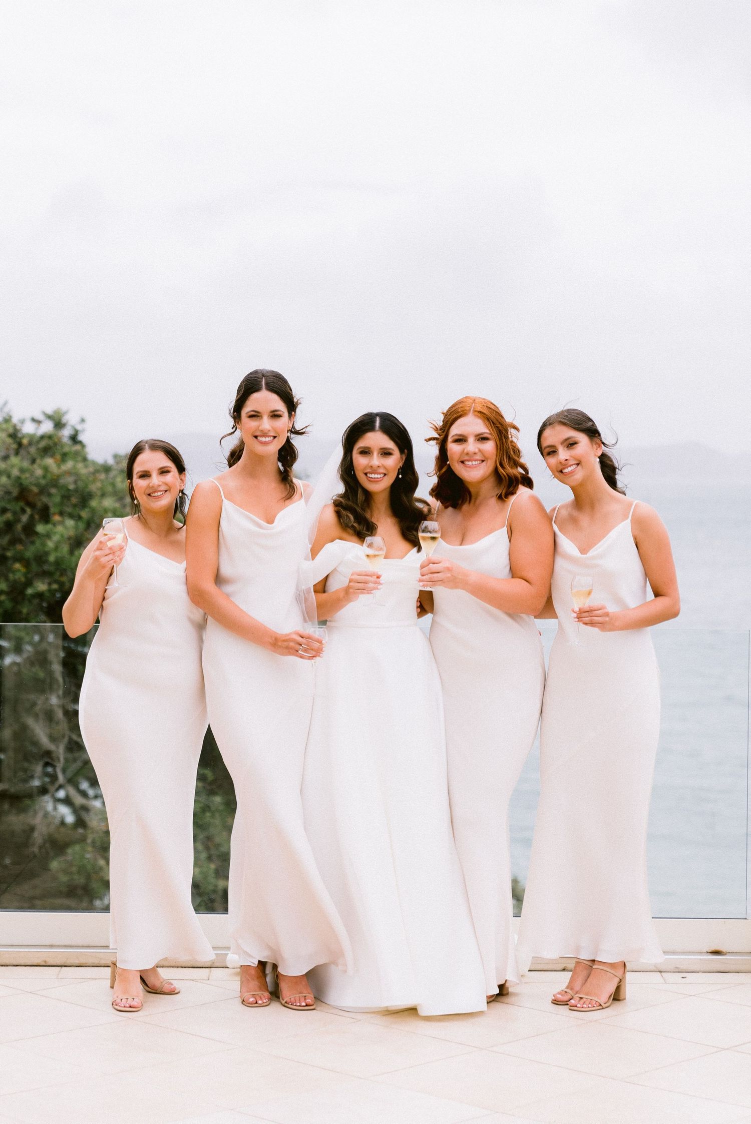 A group photo of bridesmaids wearing elegant white dresses posing together on a beachfront setting.
