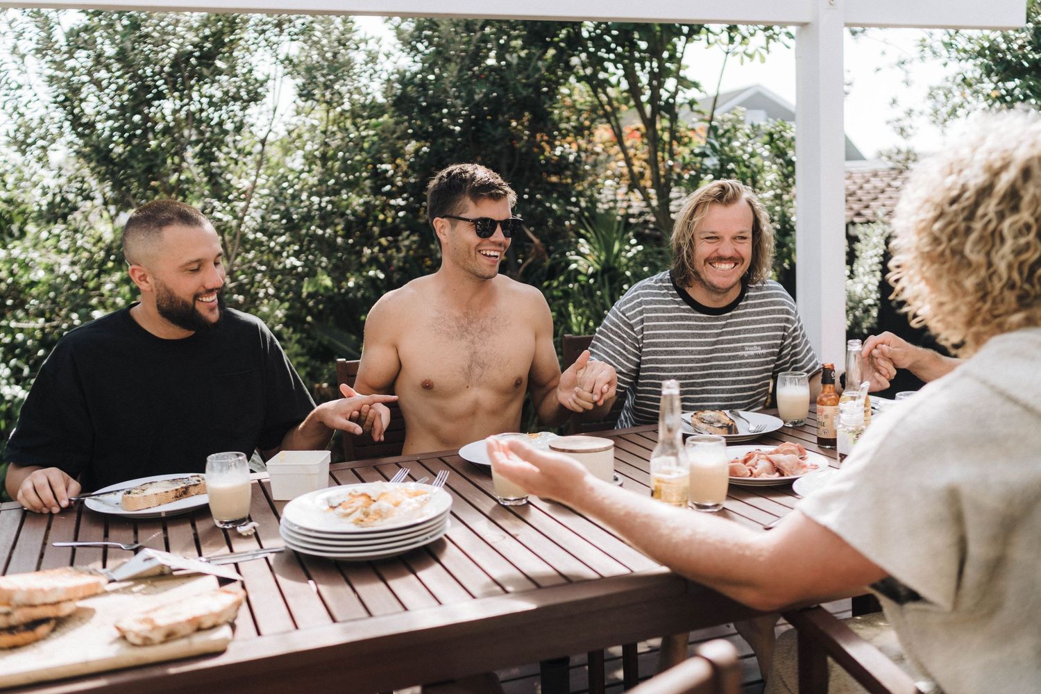 Friends gather outdoors for a casual meal at wooden tables under trees.