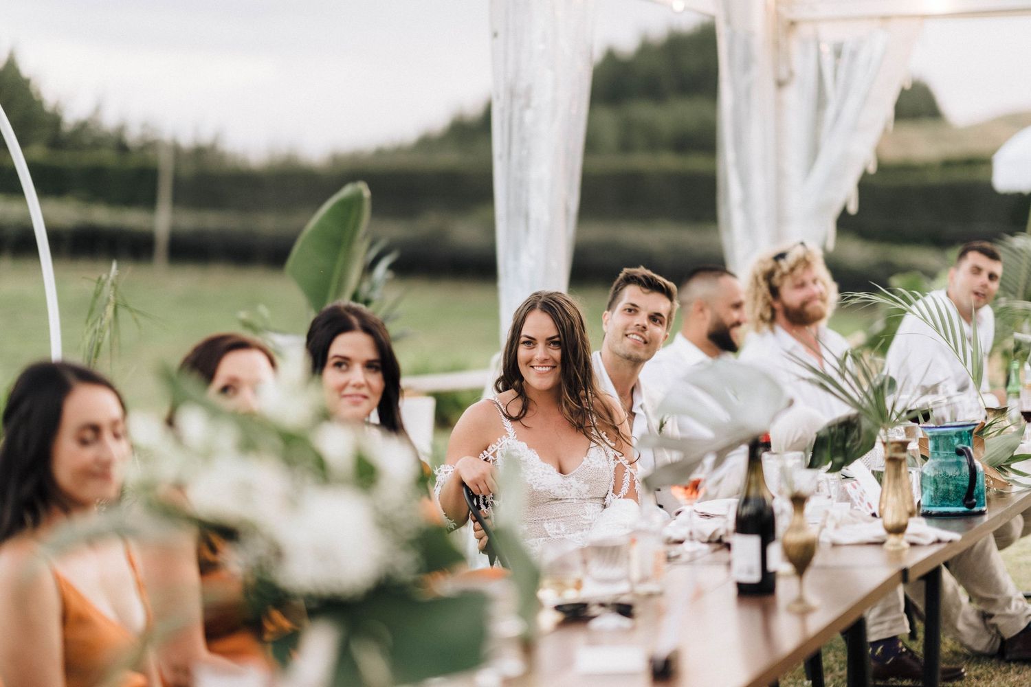 Wedding party seated at long table under outdoor tent with floral centerpieces.