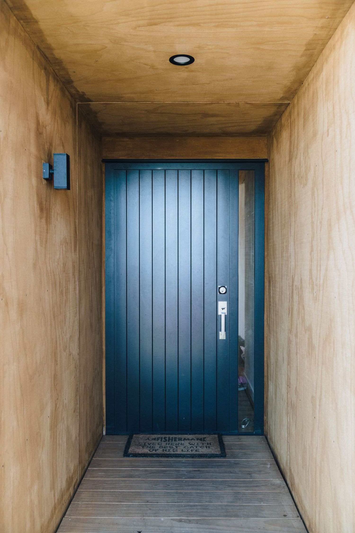 A modern blue wooden door with vertical panels at the end of a beige hallway with a welcome mat.