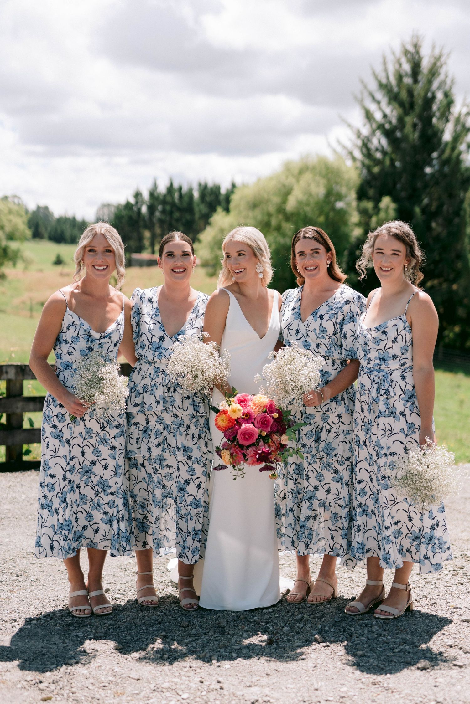 Bridesmaids in floral blue dresses pose with the bride on a sunny outdoor path.