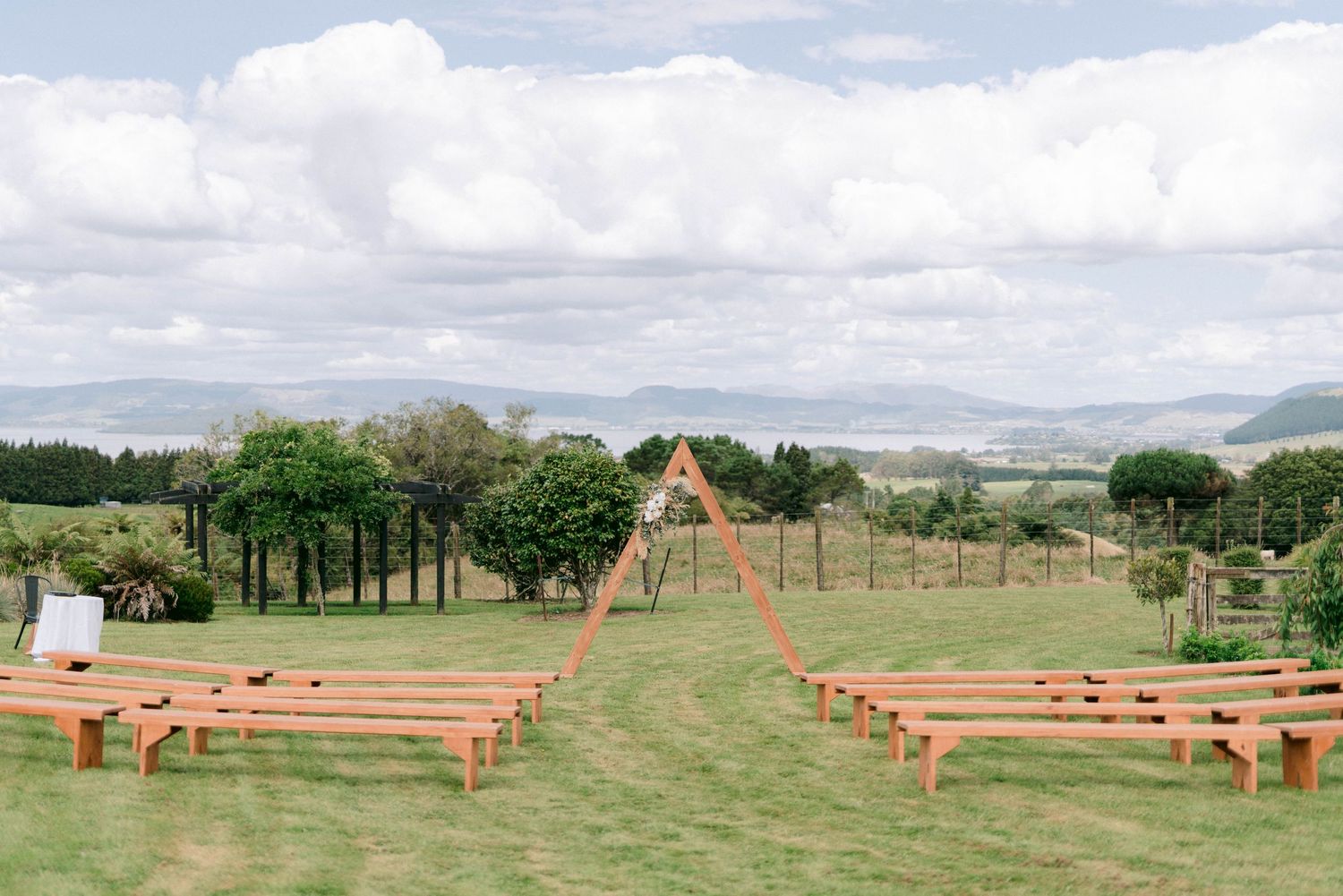 Rustic outdoor wedding ceremony setup with wooden benches facing an arch backdrop overlooking coastal scenery.