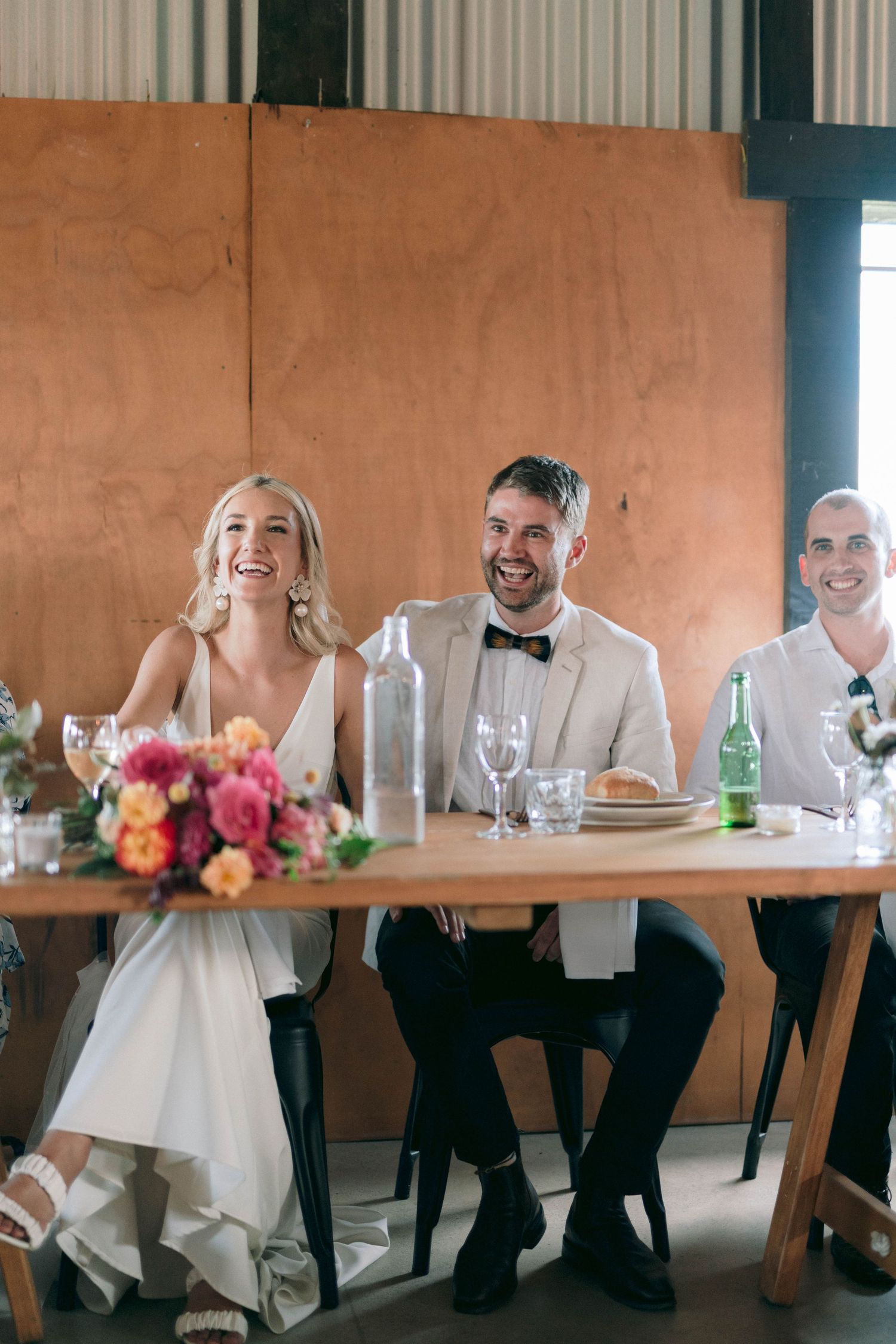 Wedding party seated at long wooden table during reception with colorful floral centerpieces.