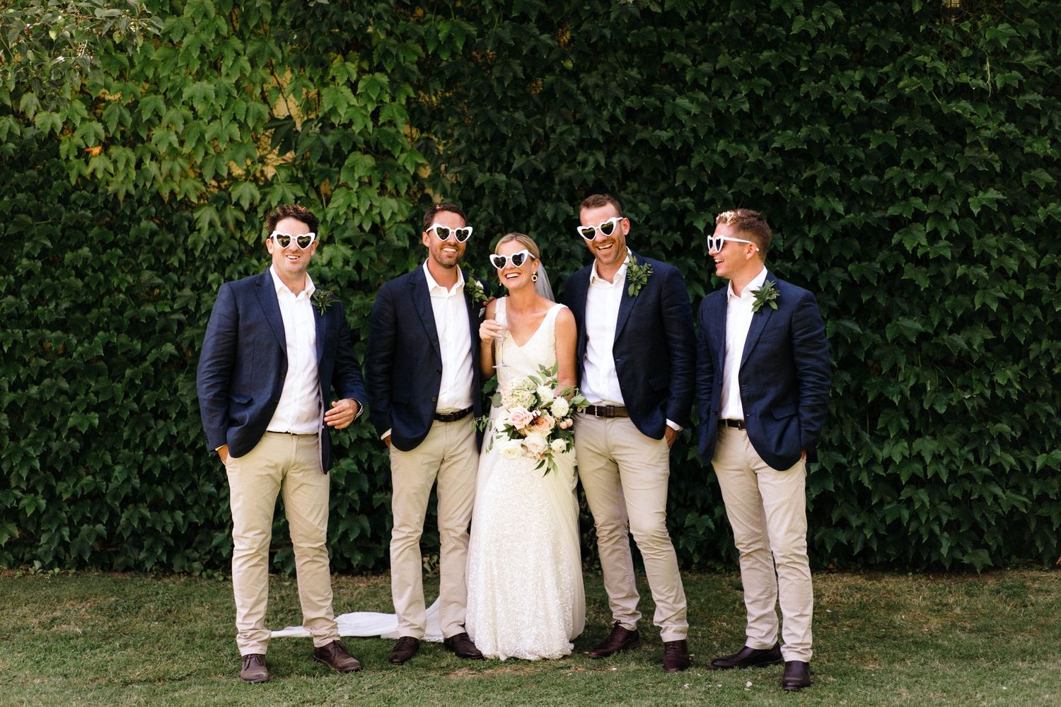 A wedding party poses together on grass against a leafy green background wearing navy and khaki attire.