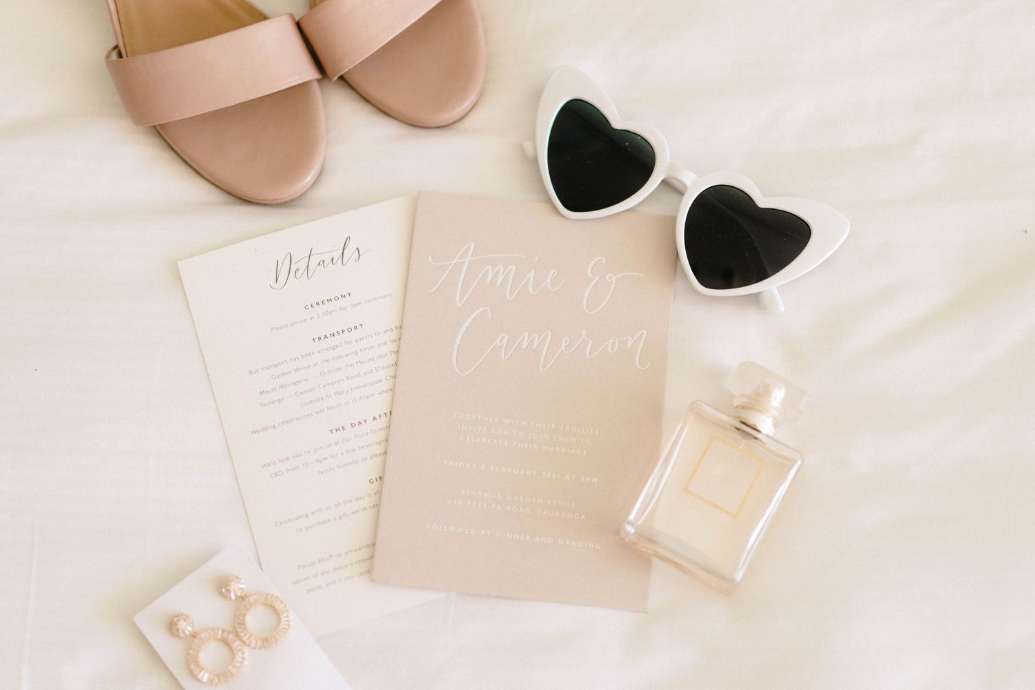 Feminine flat lay with blush sandals, heart-shaped sunglasses, perfume bottle and wedding stationery on white background.