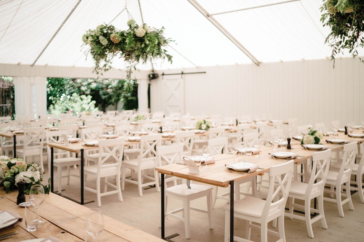 Interior view of an elegant white wedding reception tent with white chairs and wooden tables set for dining.