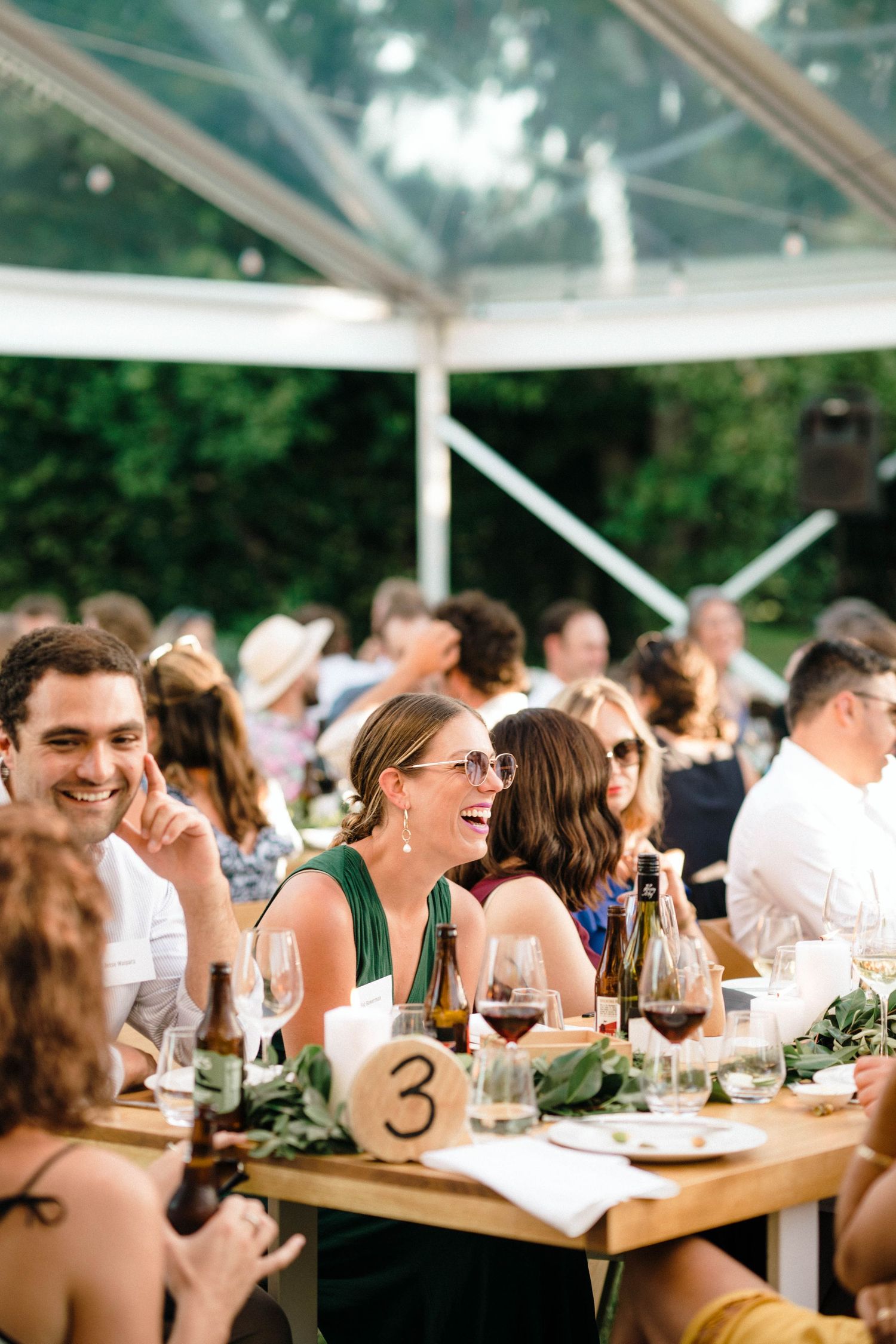 Wedding guests enjoy an elegant dinner service at long tables under a glass-roofed venue with natural lighting.