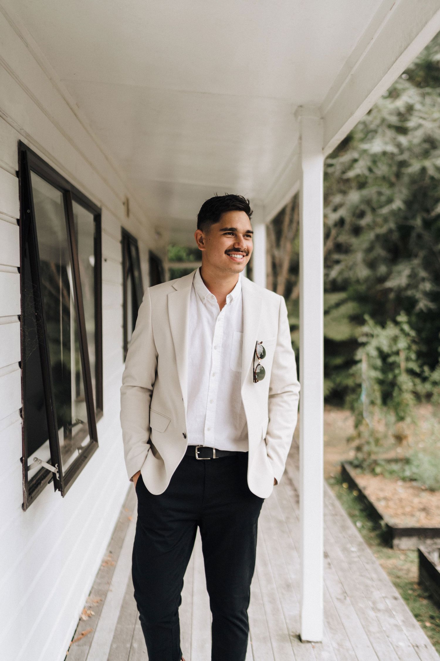 Person in white suit jacket and black pants poses on a classic white porch.