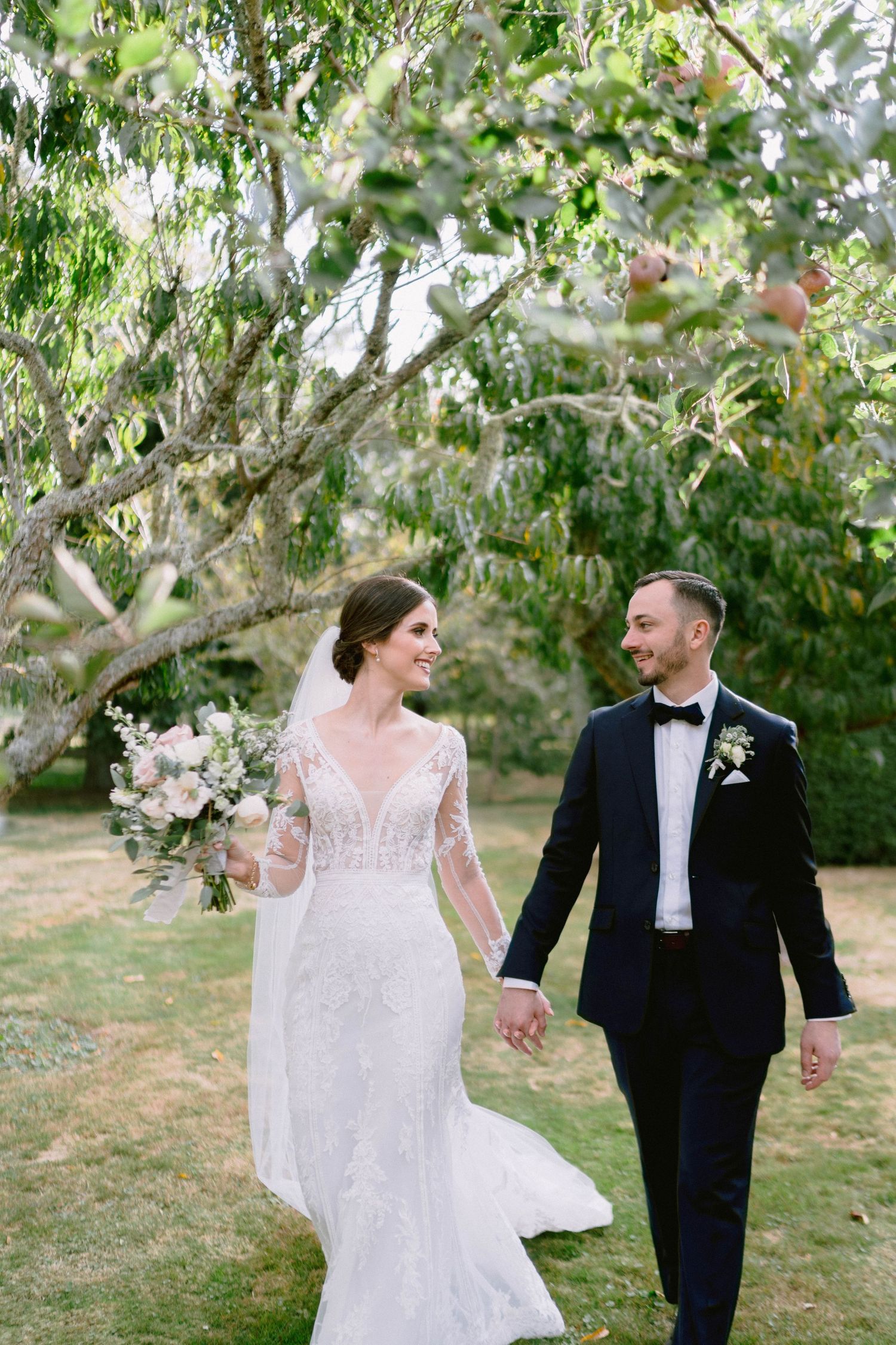 A couple walks hand in hand through an orchard on their wedding day surrounded by greenery.
