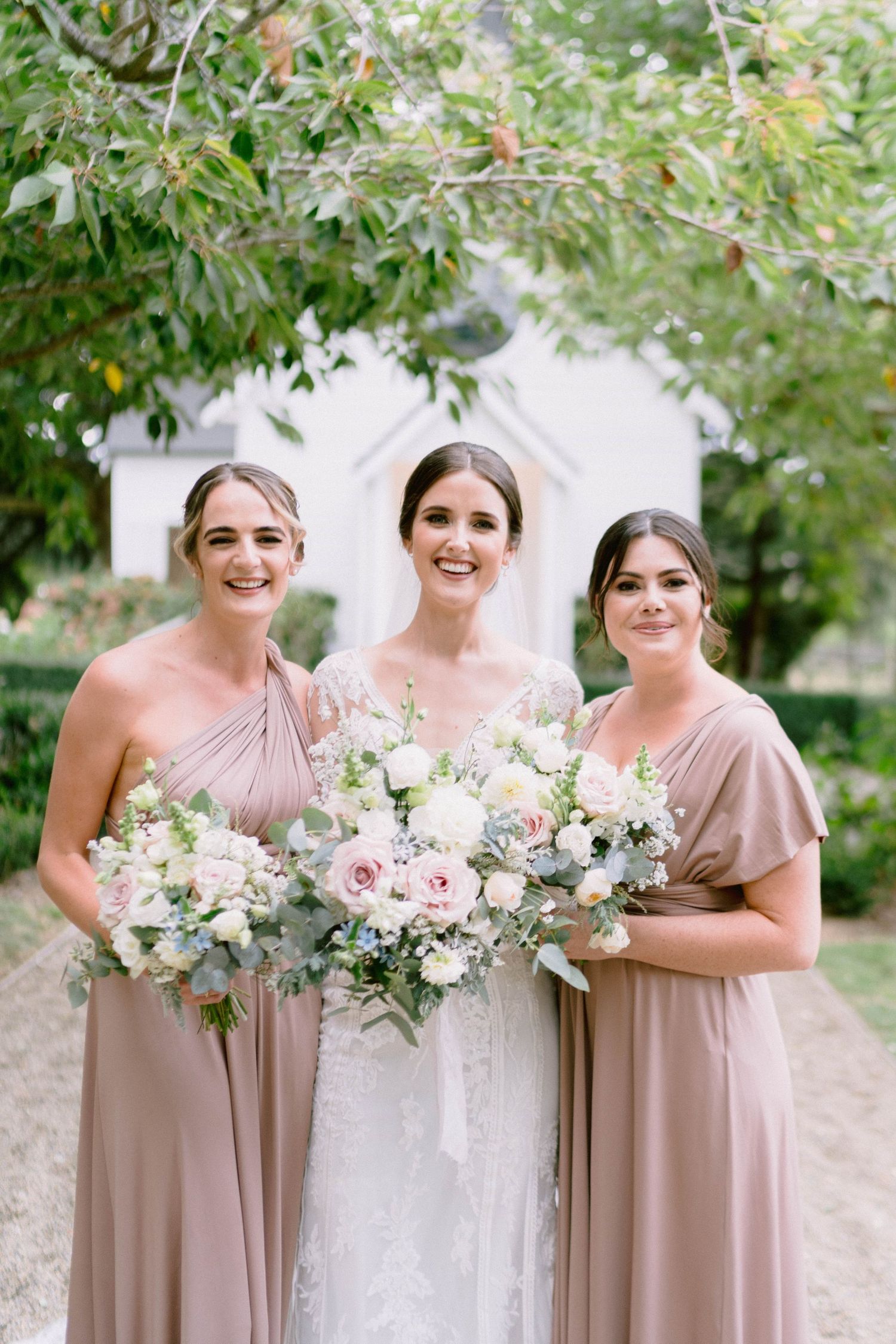 Bridesmaids in blush pink dresses hold elegant white and pink floral bouquets at an outdoor wedding.