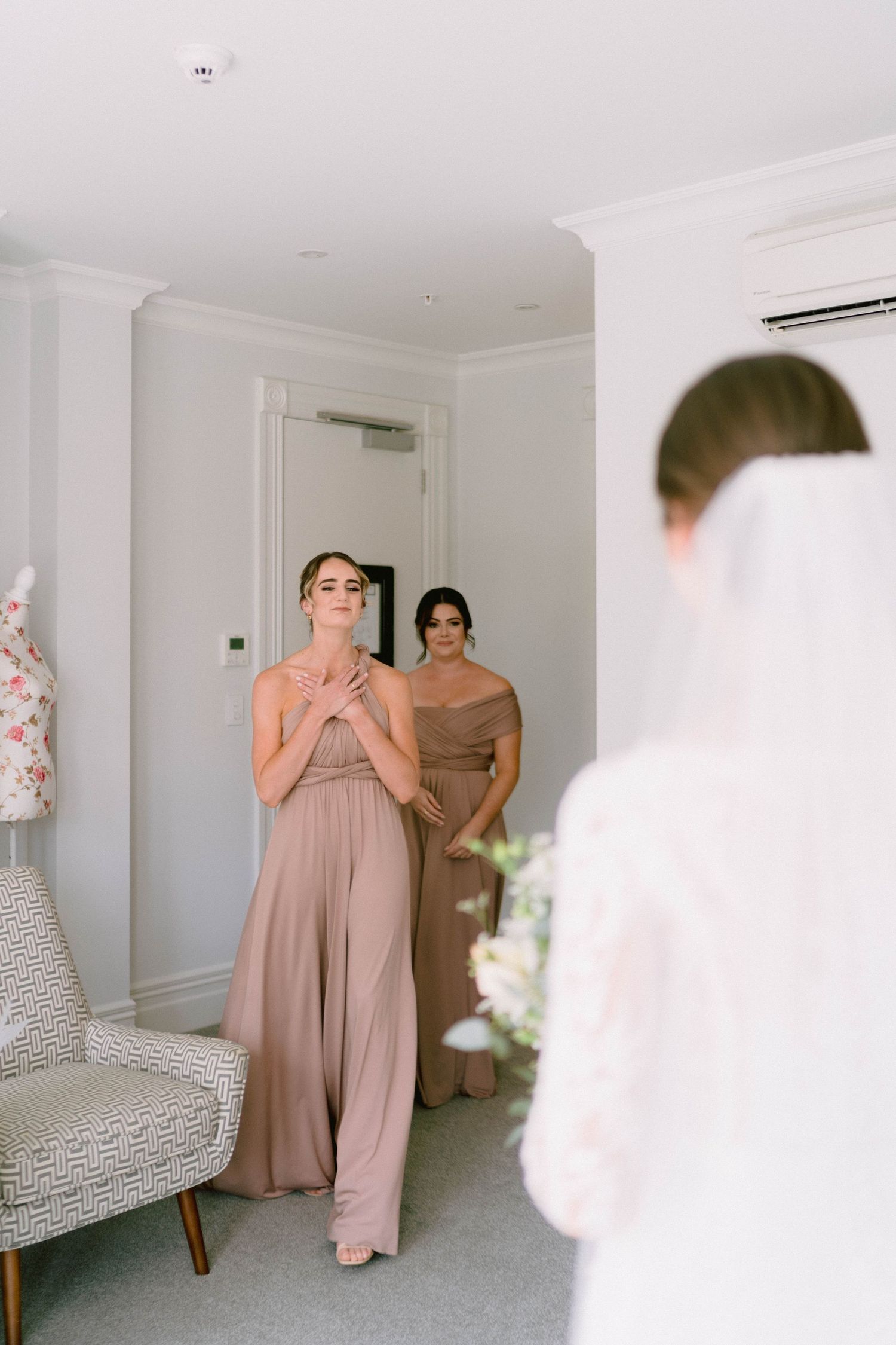 Bridesmaids in blush pink dresses share emotional reactions during wedding preparations in a bright white room.