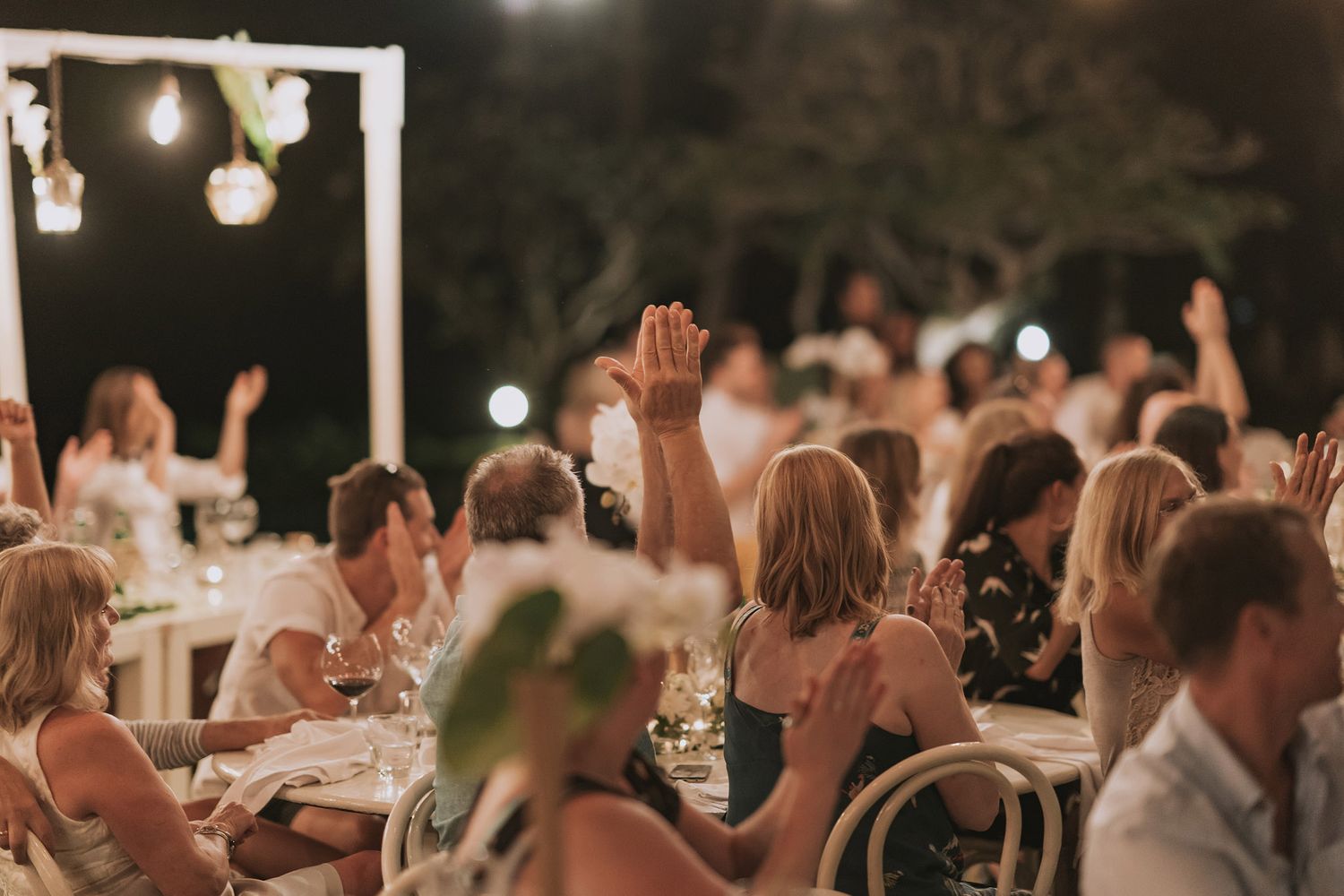 Wedding guests raise their hands in celebration during outdoor evening reception under string lights.