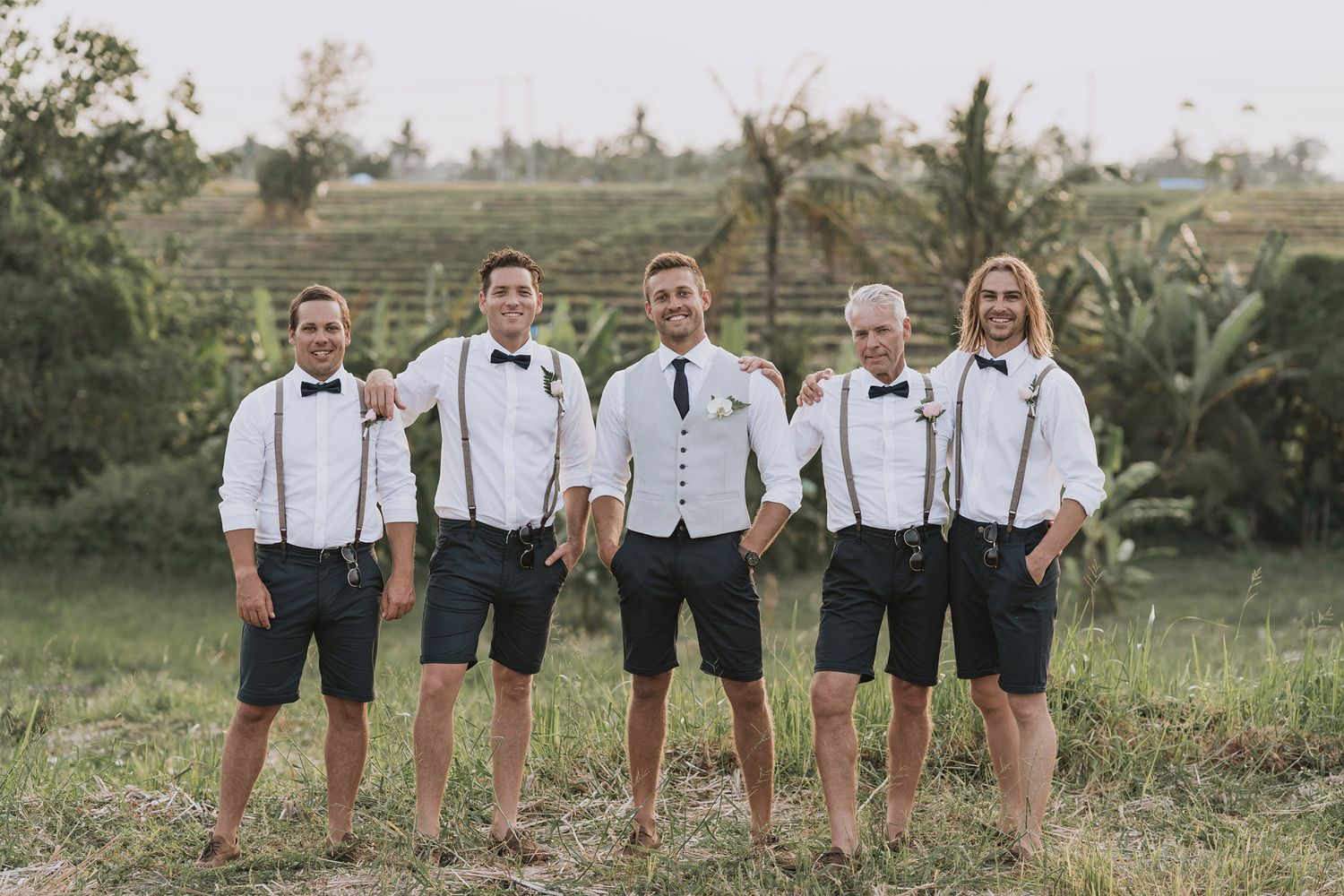 Groomsmen in matching black shorts and suspenders pose together in a tropical garden setting.