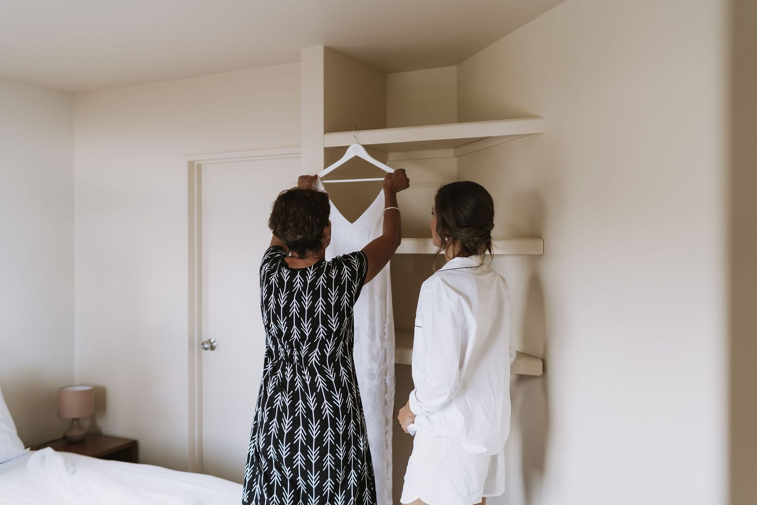 Two people organizing clothes hangers on a white closet shelf in a bright interior space.