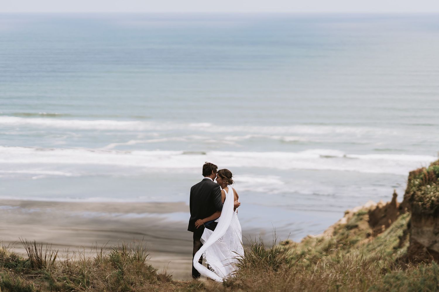Couple in wedding attire standing on coastal cliffs overlooking misty beach and ocean waves.