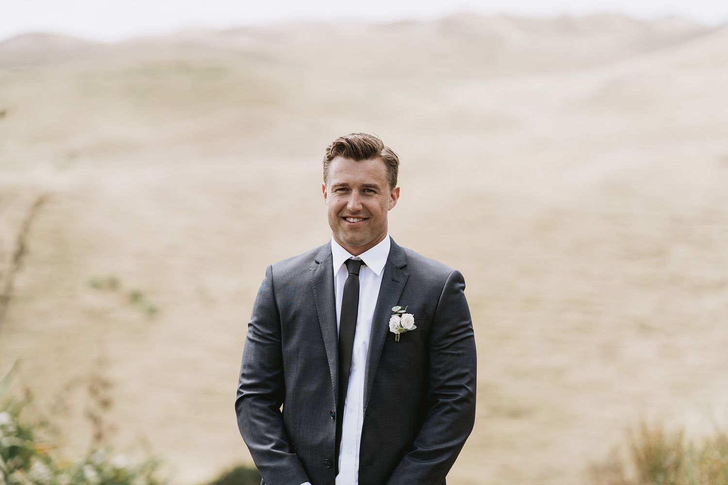 A person wearing a black suit and tie stands outdoors with a boutonniere against a blurred natural backdrop.