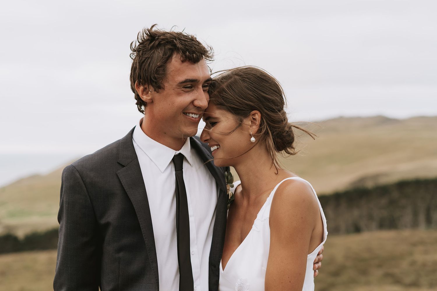 A wedding couple embraces in a golden field against rolling hills at sunset.