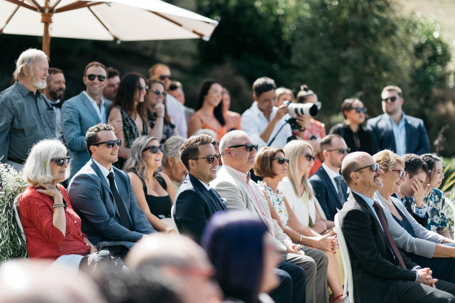 Wedding guests sit in rows of chairs outdoors under umbrellas at an elegant daytime ceremony.