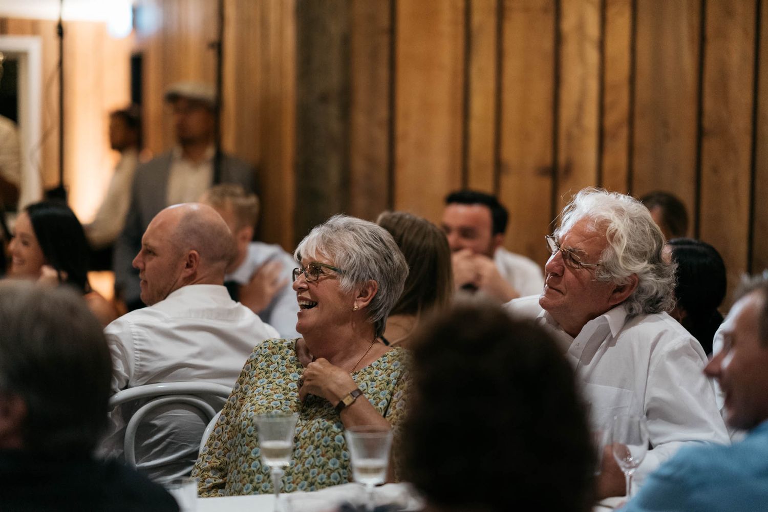Elderly guests enjoying conversation and laughter during a social gathering with wooden paneled walls in the background.
