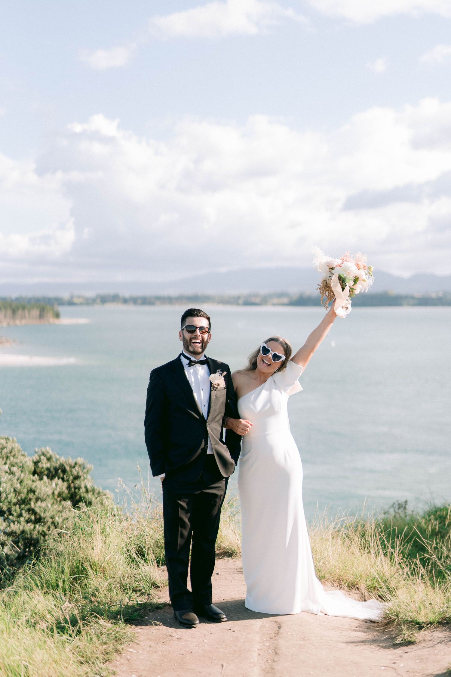 A joyful couple celebrates their wedding day on a scenic coastal clifftop with ocean views and blue skies.