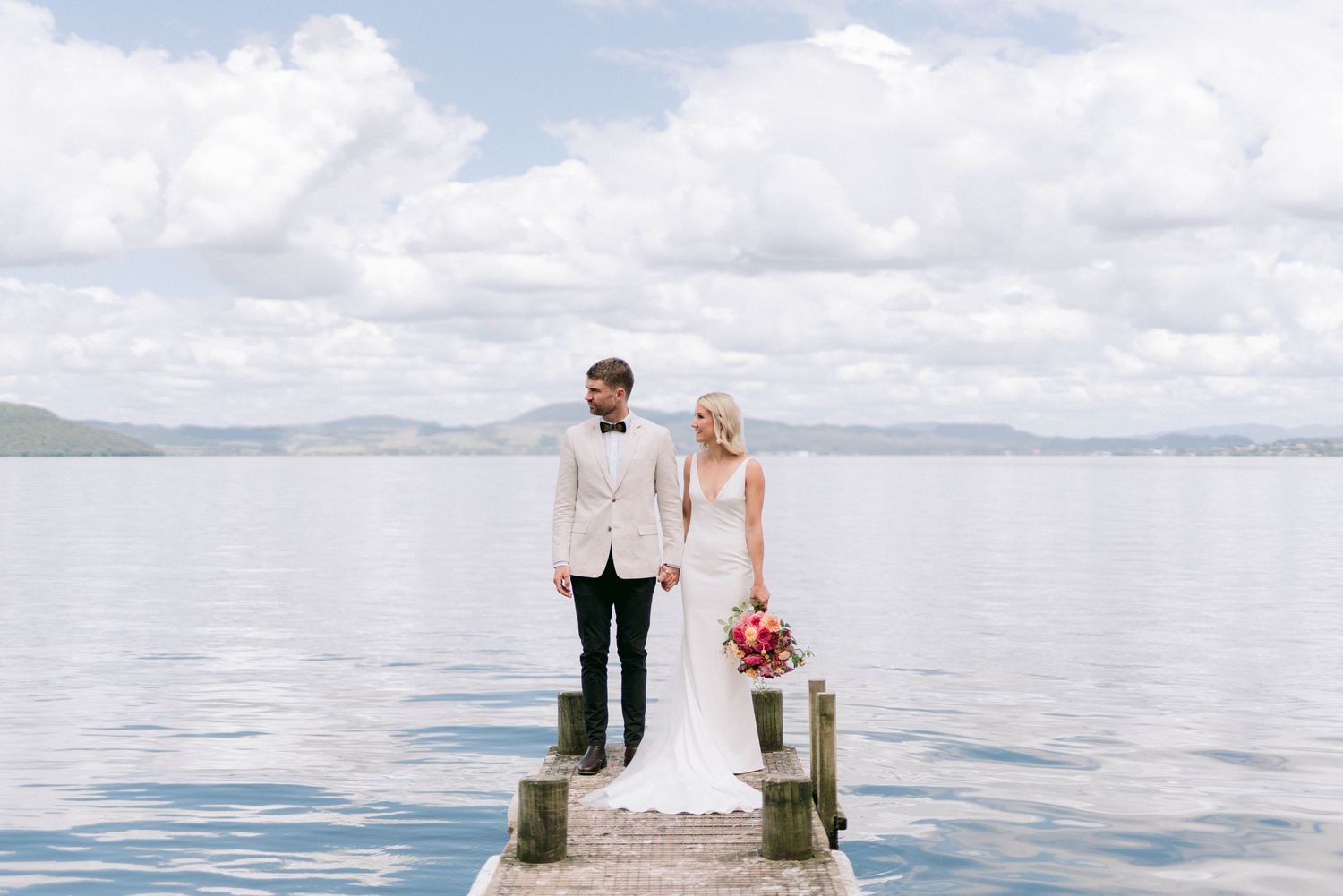 Couple stands at end of pier overlooking calm lake with mountains in background on their wedding day.