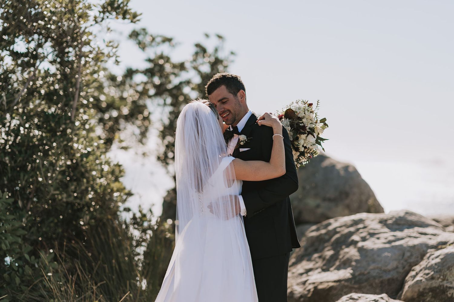 A couple in wedding attire embraces outdoors against a scenic mountain backdrop at sunset.