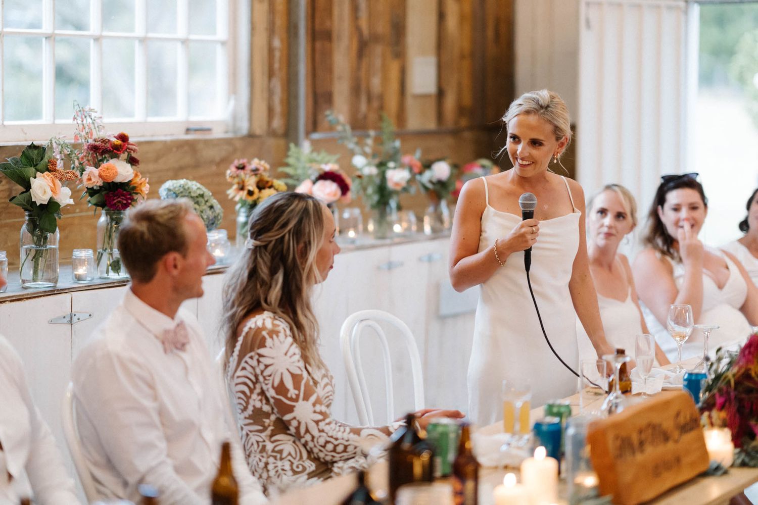 A wedding reception speech being delivered during dinner in a bright rustic-style venue with floral centerpieces.