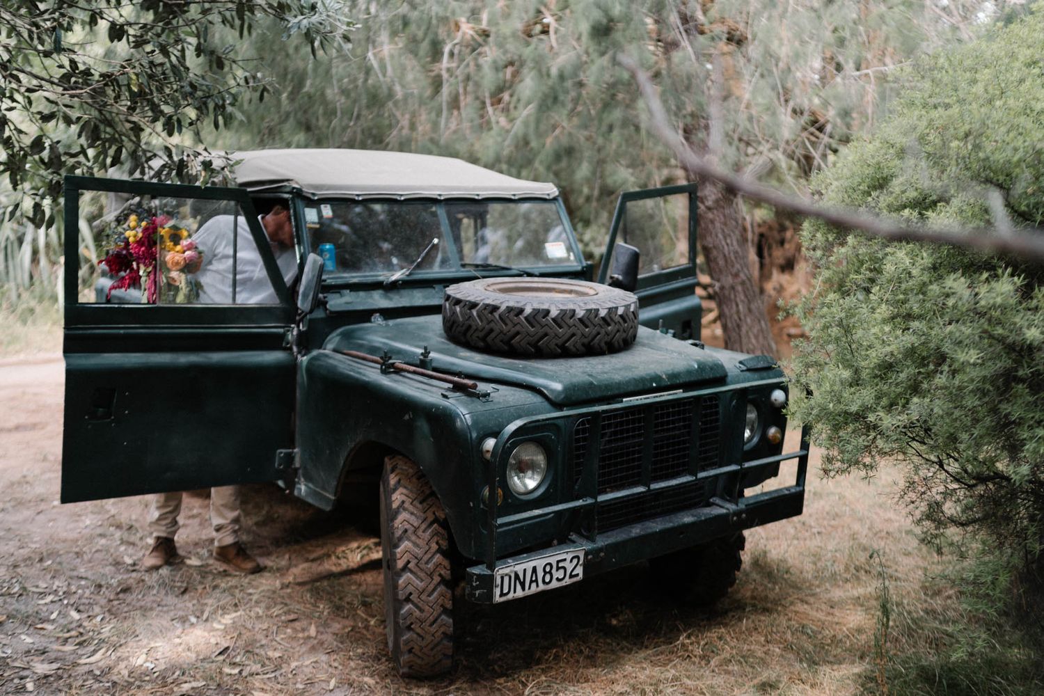 A vintage black Land Rover parked in a wooded forest setting with rugged all-terrain tires and classic styling.