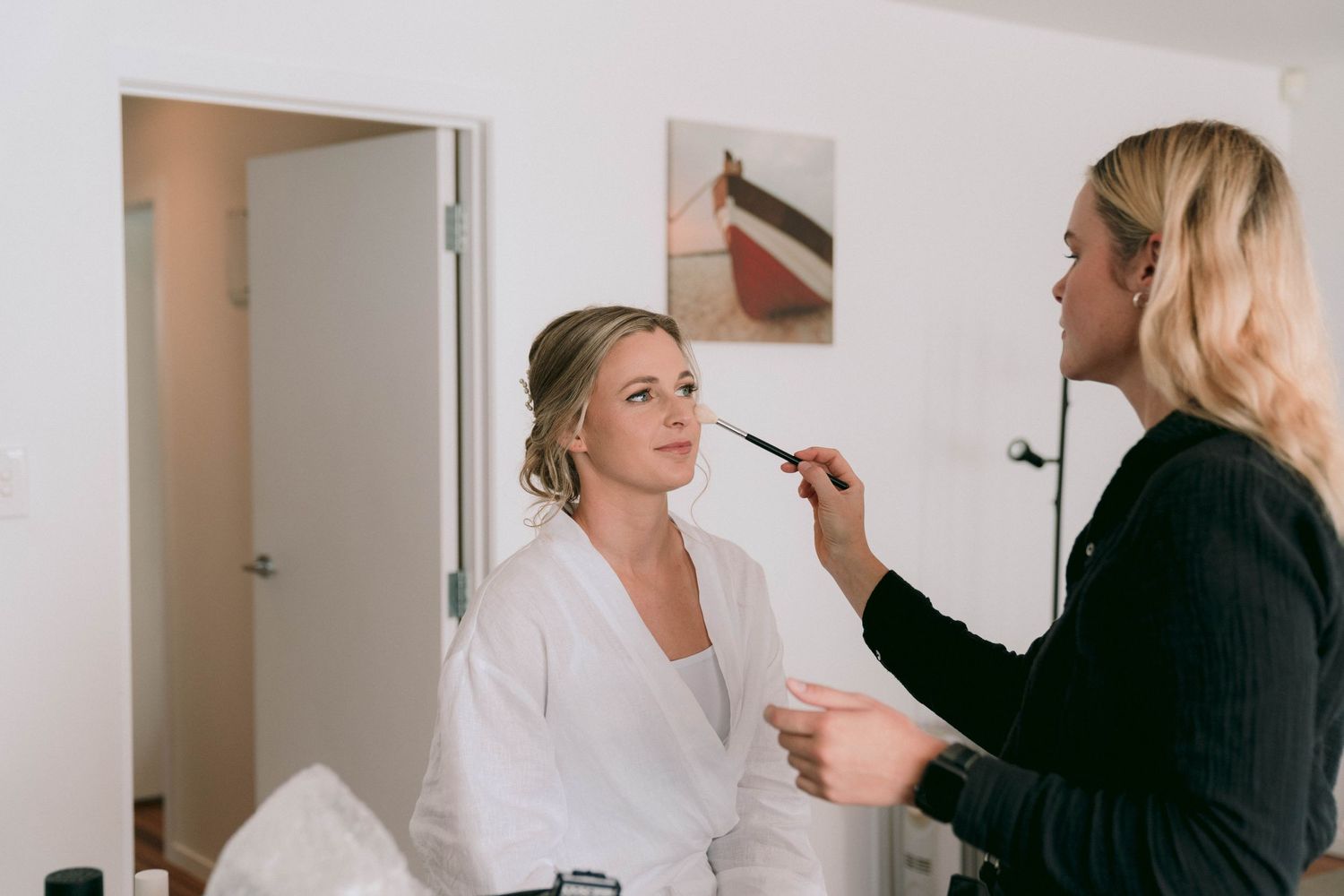 Professional makeup artist applying cosmetics to client wearing white robe in bright preparation room.