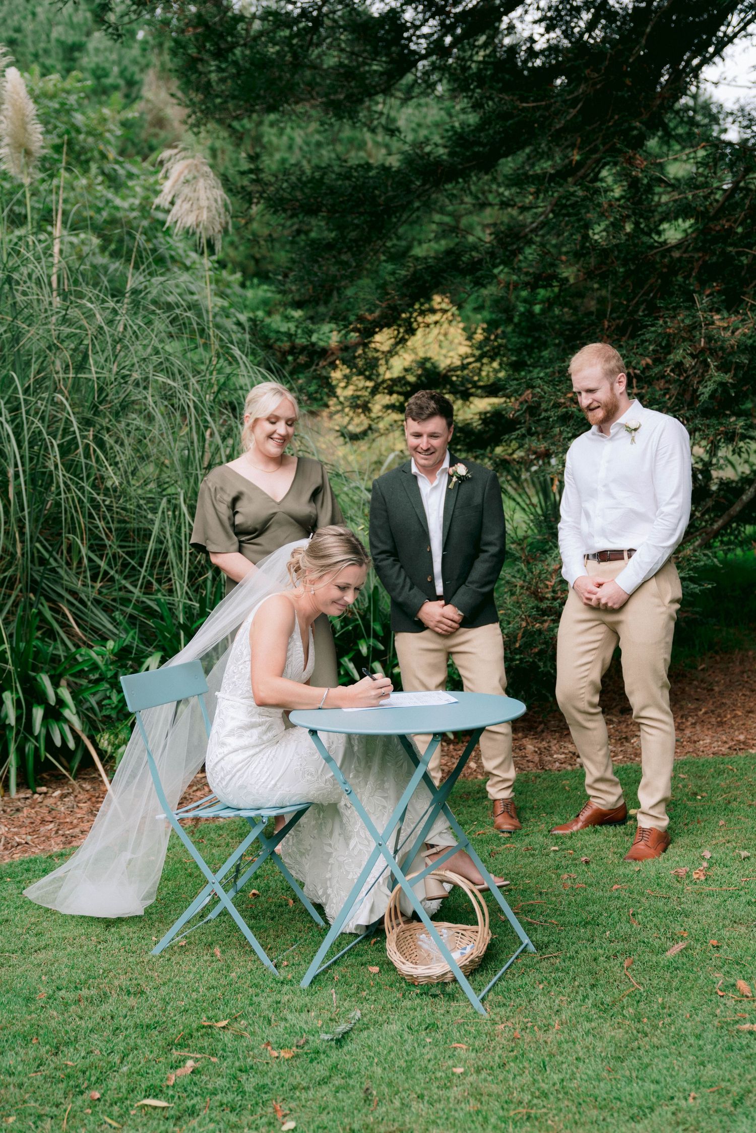 Wedding ceremony in a garden setting with a blue folding table and chairs surrounded by lush greenery.