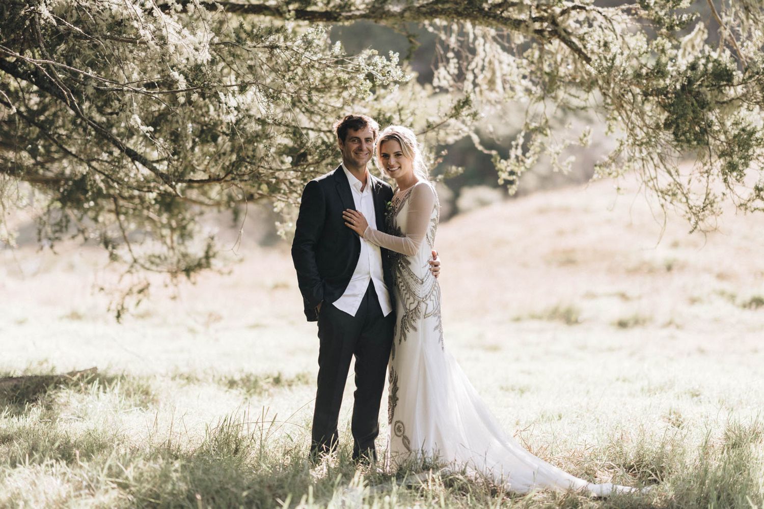 Newlyweds pose in a dreamy sunlit meadow under tree branches wearing formal wedding attire.