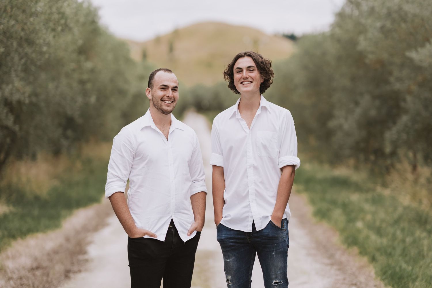 Two people walking together on a country path lined with trees and olive groves against rolling hills.