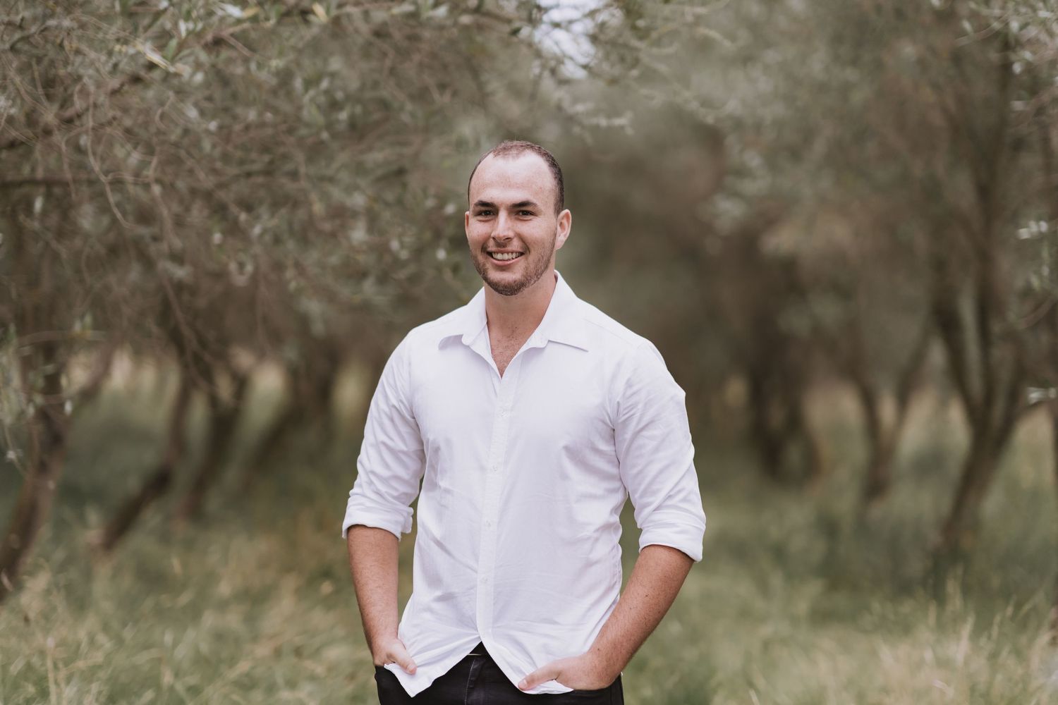 A person in a white button-down shirt stands smiling in an outdoor setting with trees in the background.