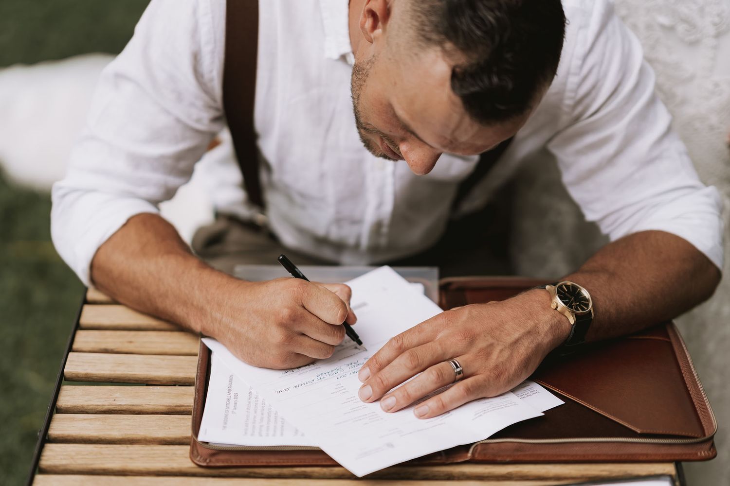Person wearing white dress shirt writing on paper at wooden table with brown notebook outdoors.