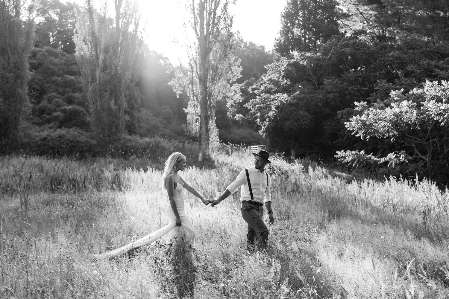 A romantic black and white photo shows a couple holding hands while standing in a sunlit meadow surrounded by trees.